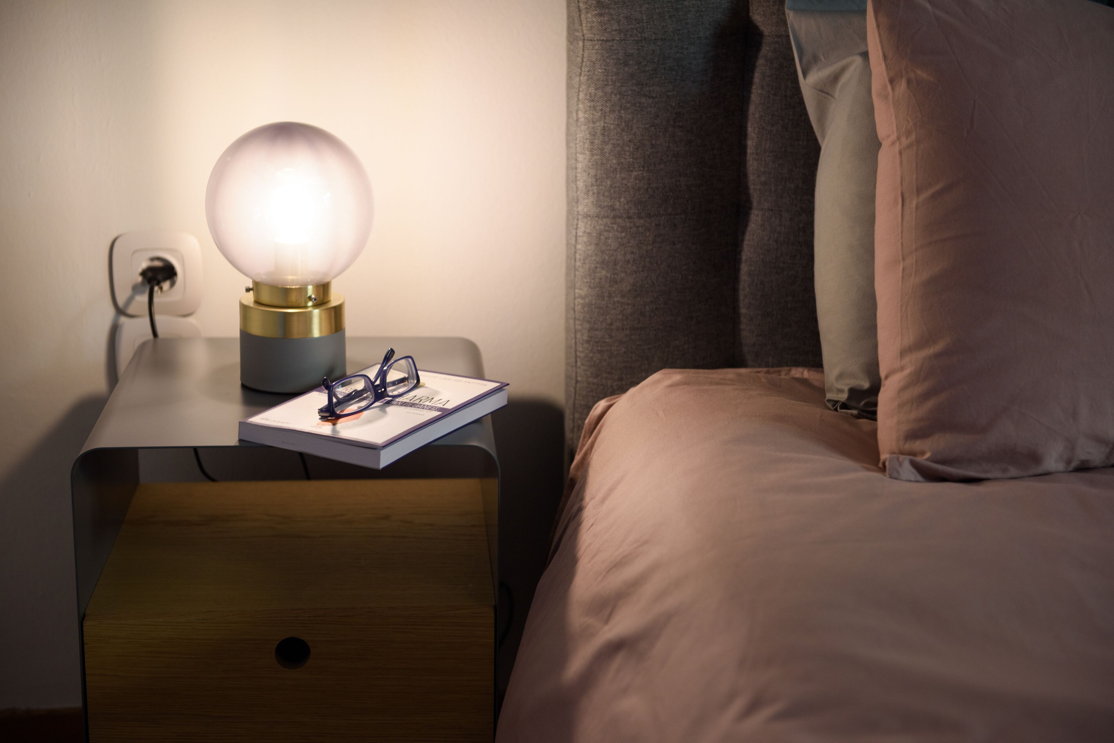 Un lit bien fait, avec une tête de lit, une table de chevet sur laquelle se trouve une lampe, et une paire de lunettes posée sur un carnet.