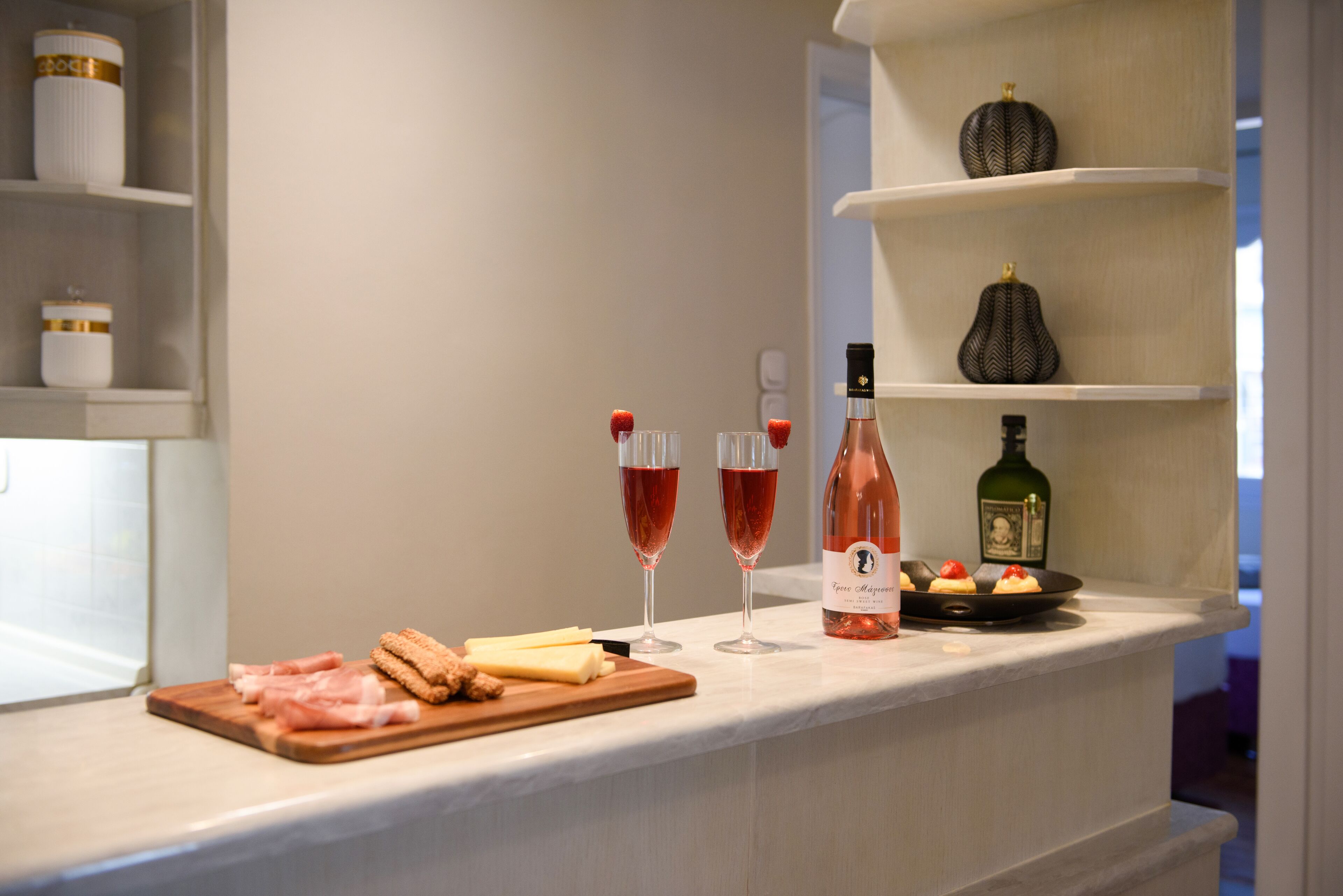A kitchen counter with a cutting board, sliced meats, cheese, and two glasses of rosé wine.