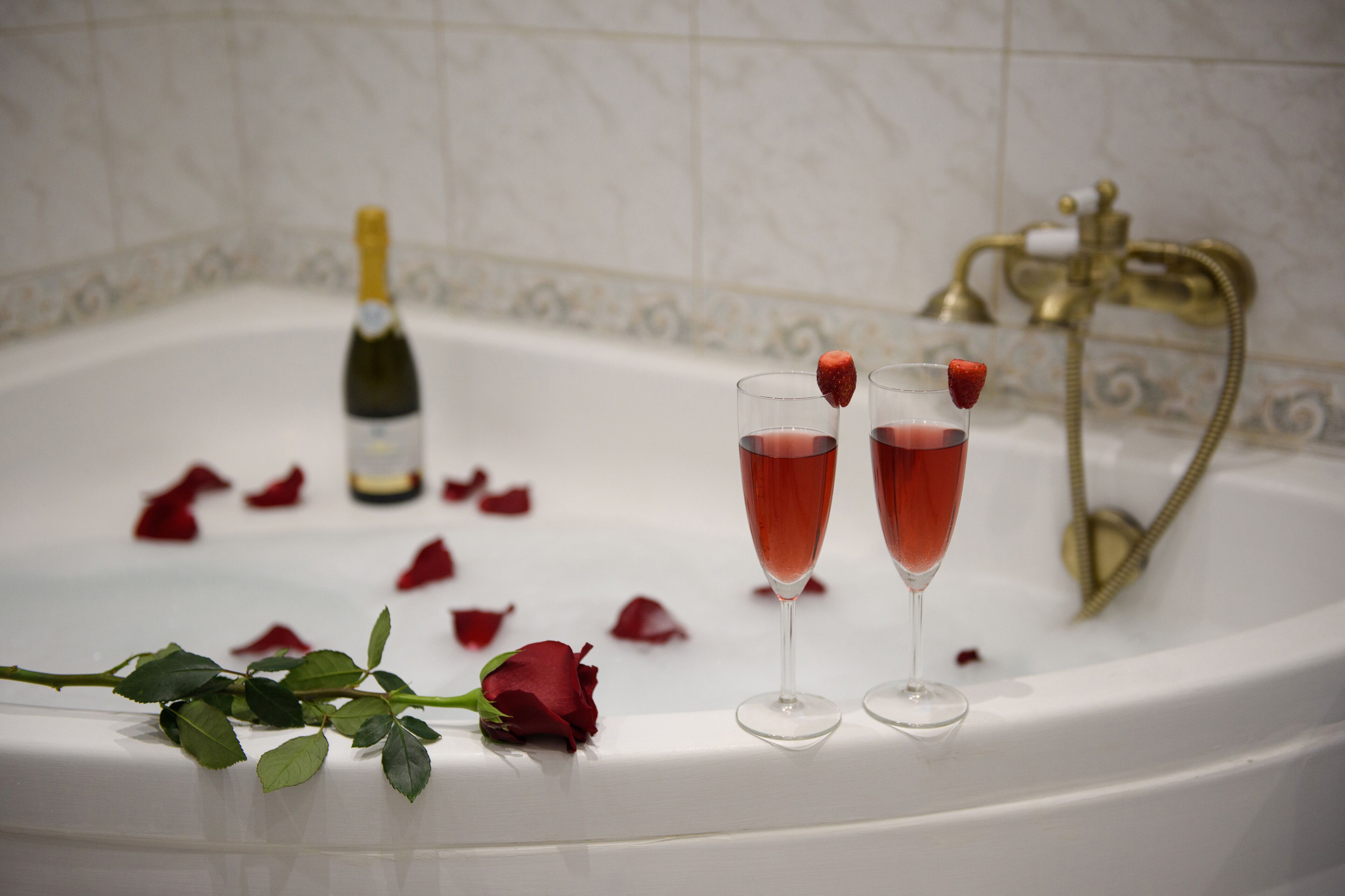A bathtub with champagne and rose petals, a rose, and a gold faucet.