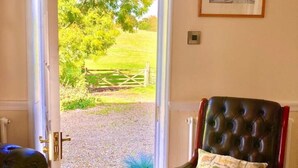 A room with a view of a grassy field through a glass door, a leather armchair with a floral cushion, and a framed picture on the wall.