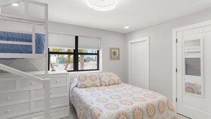 A bedroom with a bunk bed, a bed with a patterned bedspread, a window with blinds, a door, and a ceiling fan.