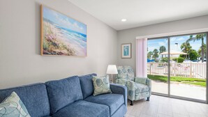 A living room with a blue sofa, a chair, a painting of a beach, and a view of a parking area with palm trees.