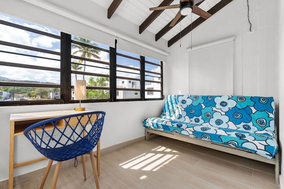 Une pièce comprenant un bureau, une chaise bleue, un canapé à motifs et une grande fenêtre donnant sur des palmiers et des immeubles.