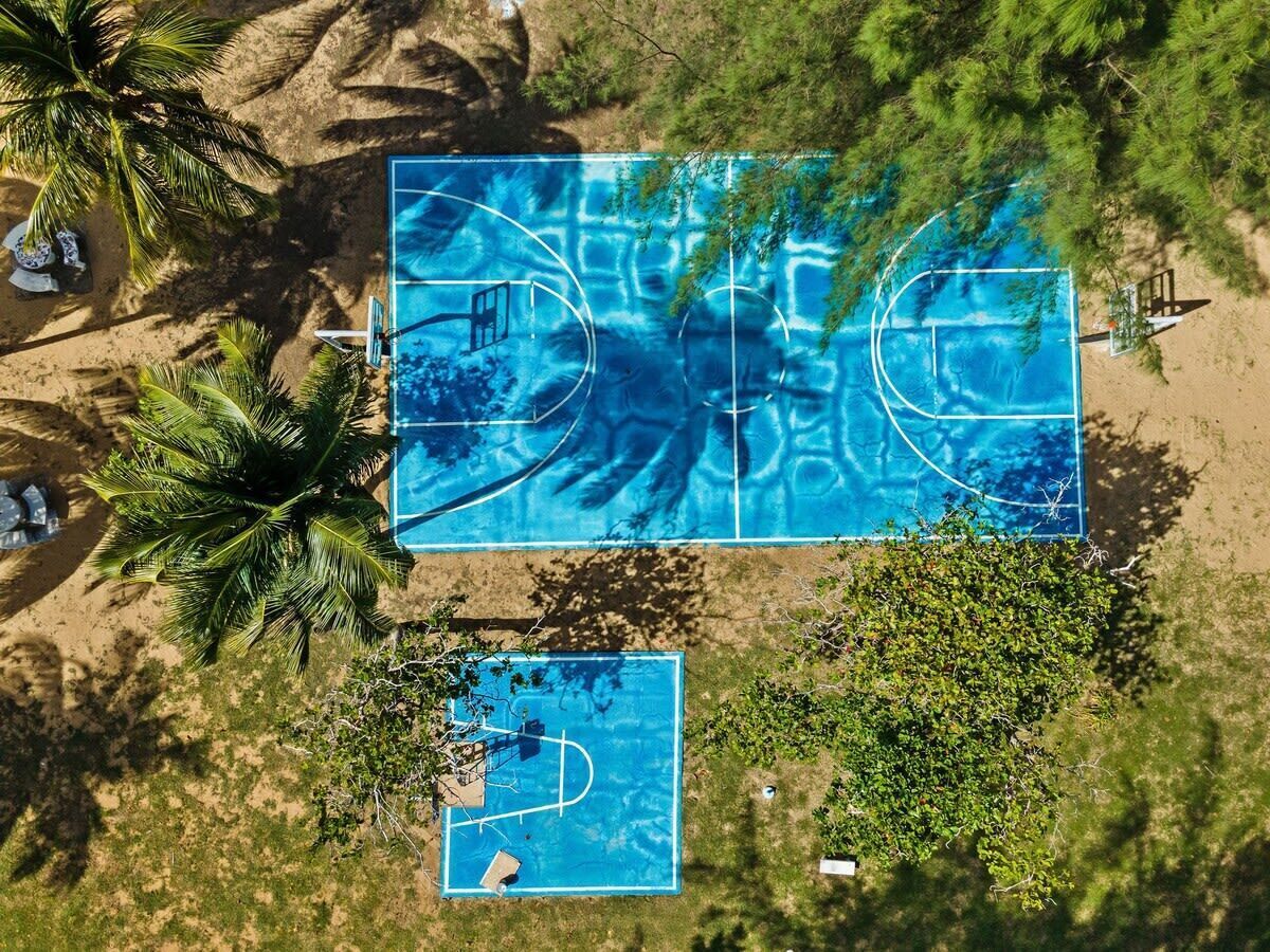An aerial view of a beach with two blue basketball courts surrounded by palm trees and greenery.