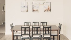 A dining area with a long wooden table, six chairs, and three framed pictures on the wall. - Brand New Ski in/out + Hot Tub!. (Big White)