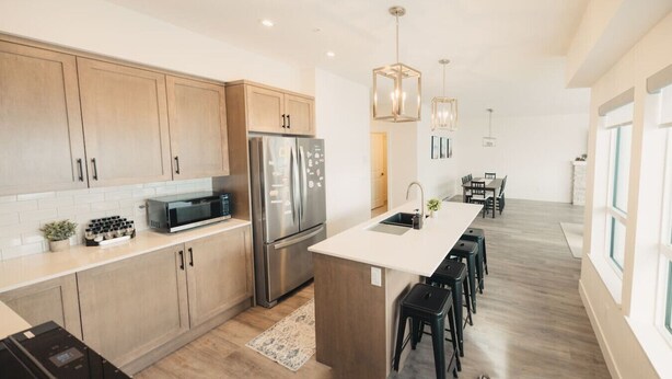 A modern kitchen with wooden cabinets, a stainless steel refrigerator, a microwave, and a dining area with a table and chairs. - Brand New Ski in/out + Hot Tub!. (Big White)