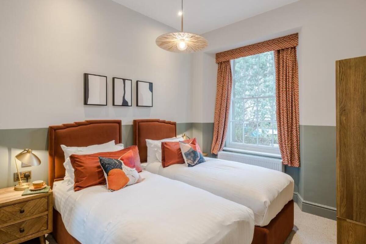 Habitación doble con dos camas, una mesita de noche, una lámpara y un ventanal con cortinas estampadas.
