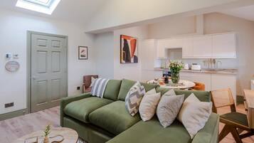 Una sala de estar moderna con un sofá verde, una mesa de centro redonda y una cocina con armarios blancos.