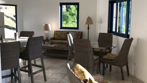 Una habitación amplia con techo alto, ventilador de techo, comedor con mobiliario de mimbre y ventanas grandes que ofrecen vista a la vegetación.