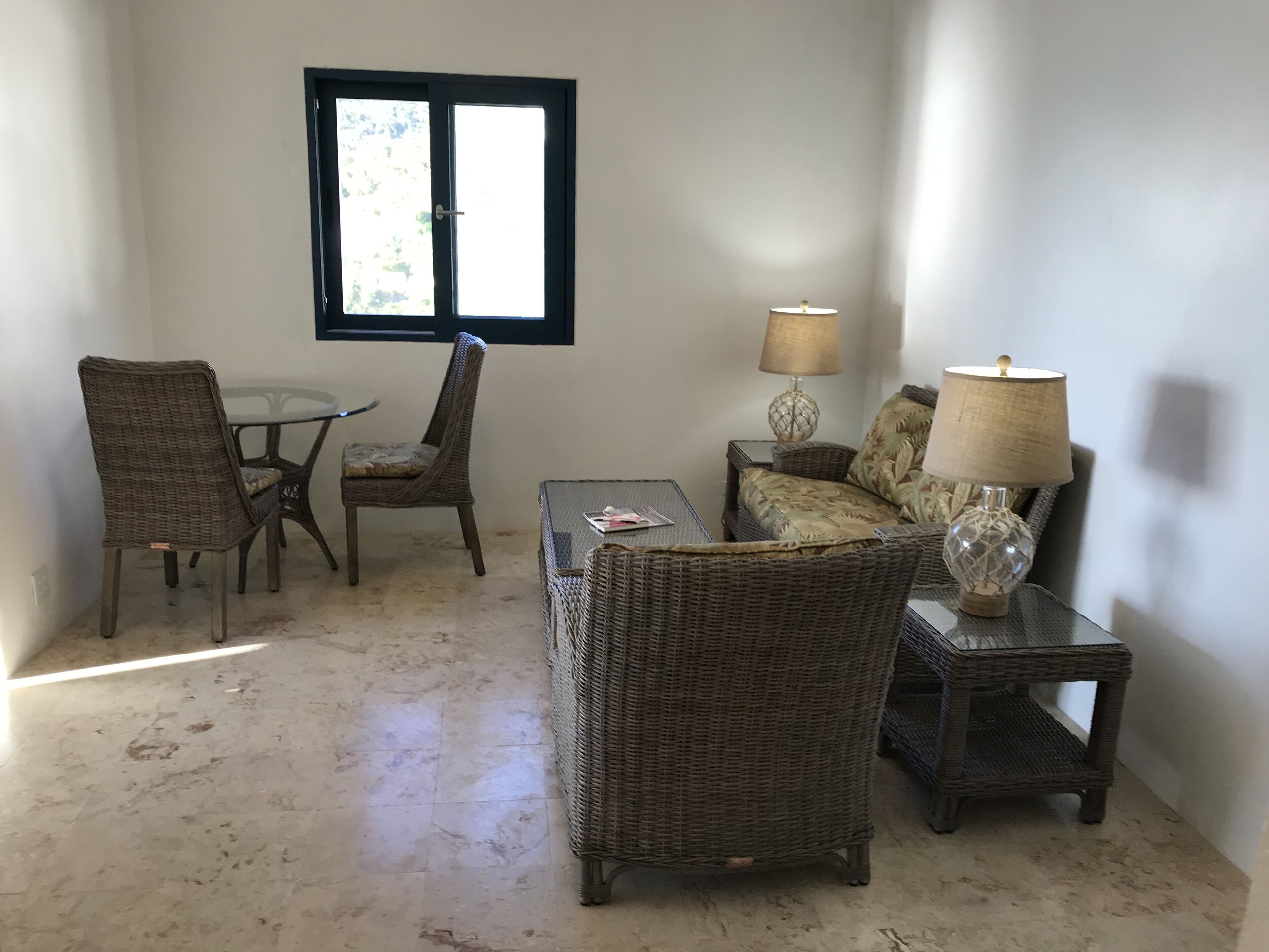 Una habitación con muebles de mimbre, una mesa de vidrio, un sofá y dos lámparas.