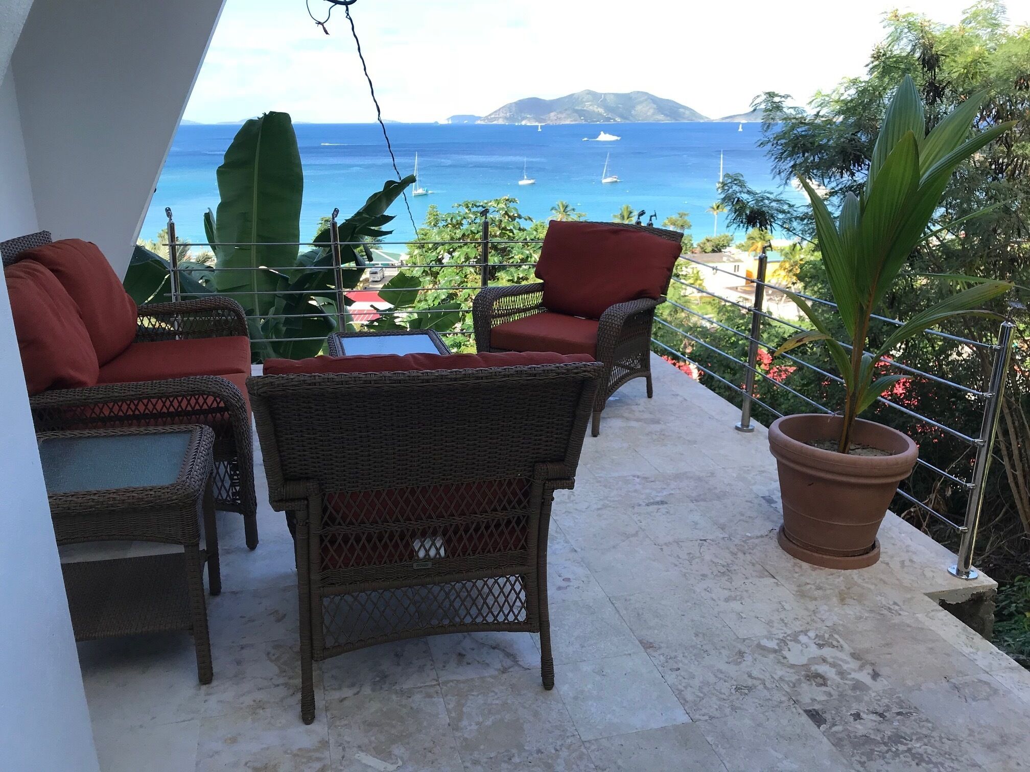 A balcony with wicker furniture, a potted plant, and a view of the sea with boats.