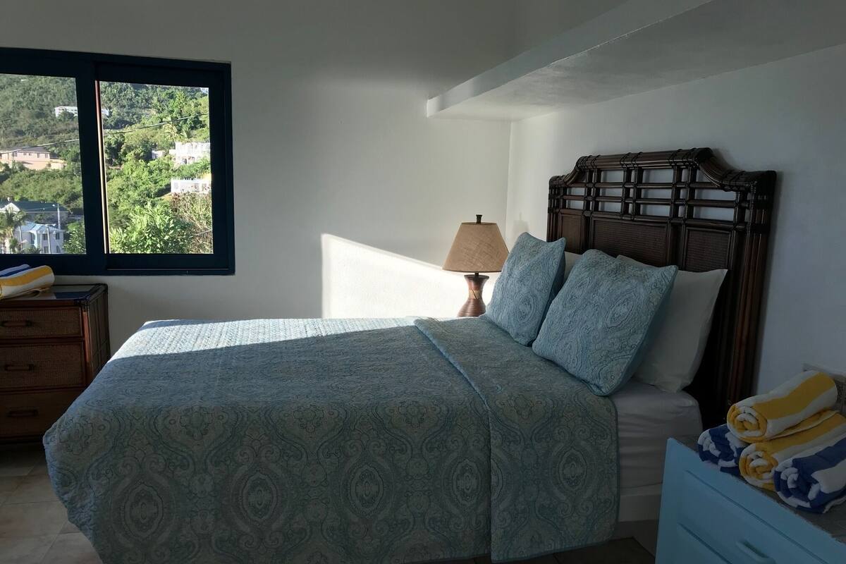 Un dormitorio con cama, mesita de noche, lámpara y vista al exterior.