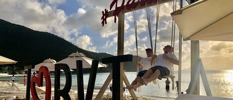 A beach with a swing set and umbrellas, a sign reading "Paradise", and a clear view of the water.