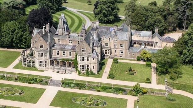 Une vue aérienne d’un vaste manoir historique doté de plusieurs ailes, entouré de pelouses parfaitement entretenues et d’un terrain de golf.