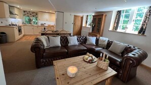 Un salon chaleureux avec un canapé en cuir marron, une table basse en bois sur laquelle est posée une bougie, et une cuisine en arrière-plan.