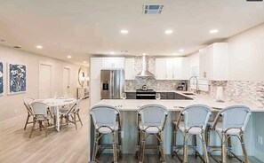 A modern kitchen with white cabinets, a central island with bar stools, and a dining area with a round table and chairs. - 668 Tres Fuentes · Heated Designer Pool w/ Spa (Naples)