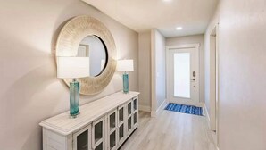 Un couloir avec un miroir rond, deux lampes, une console blanche et un tapis bleu à l’entrée.