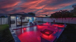 Un área de piscina moderna al aire libre con un patio de cristal, iluminación roja y vista a una casa.