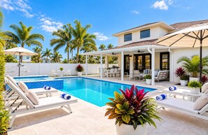 A swimming pool with lounge chairs, a covered patio area, and tropical plants.