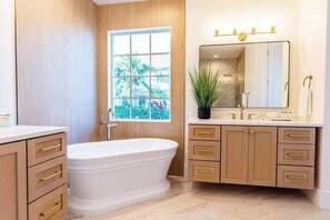 A bathroom with a freestanding bathtub, wooden cabinetry, a large mirror, and a window with a view of greenery.