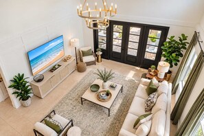 A spacious living room with a large flat-screen TV, a comfortable sofa set, a coffee table, and a dining area with a chandelier.
