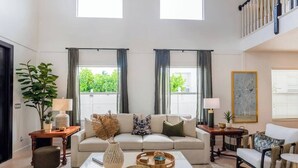 Ein geräumiges Wohnzimmer mit Sofa, Couchtisch und Beistelltischen, großen Fenstern mit Vorhängen und einer Treppe.