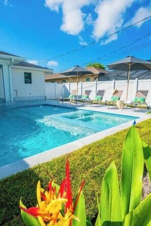 A swimming pool with lounge chairs, a white building, and tropical plants. - 5331 Aqua Haven - Waterfall Pool (Naples)
