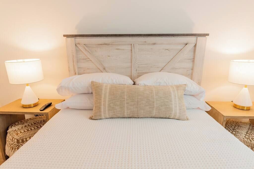 Ein Bett mit Holz-Kopfteil, zwei Nachttischlampen, einer Fernbedienung und einem gemusterten Zierkissen.