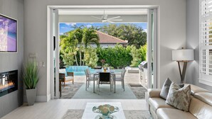 Ein modernes Wohnzimmer mit Kamin, einer Essecke mit Tisch und Stühlen, einer Couch und Blick auf die Terrasse mit Pool und Grünflächen.