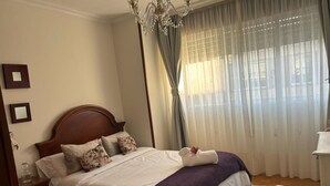 Una cama bien hecha con cabecera de madera, una manta morada y almohadas con motivos florales. Se observa una mesita de noche con un vaso y un libro. Una silla es visible en la esquina, y un candelabro cuelga del techo.