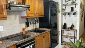 Una cocina con armarios de madera, un refrigerador negro, una estufa y un lavamanos. Hay utensilios colgando y una cafetera en los estantes.