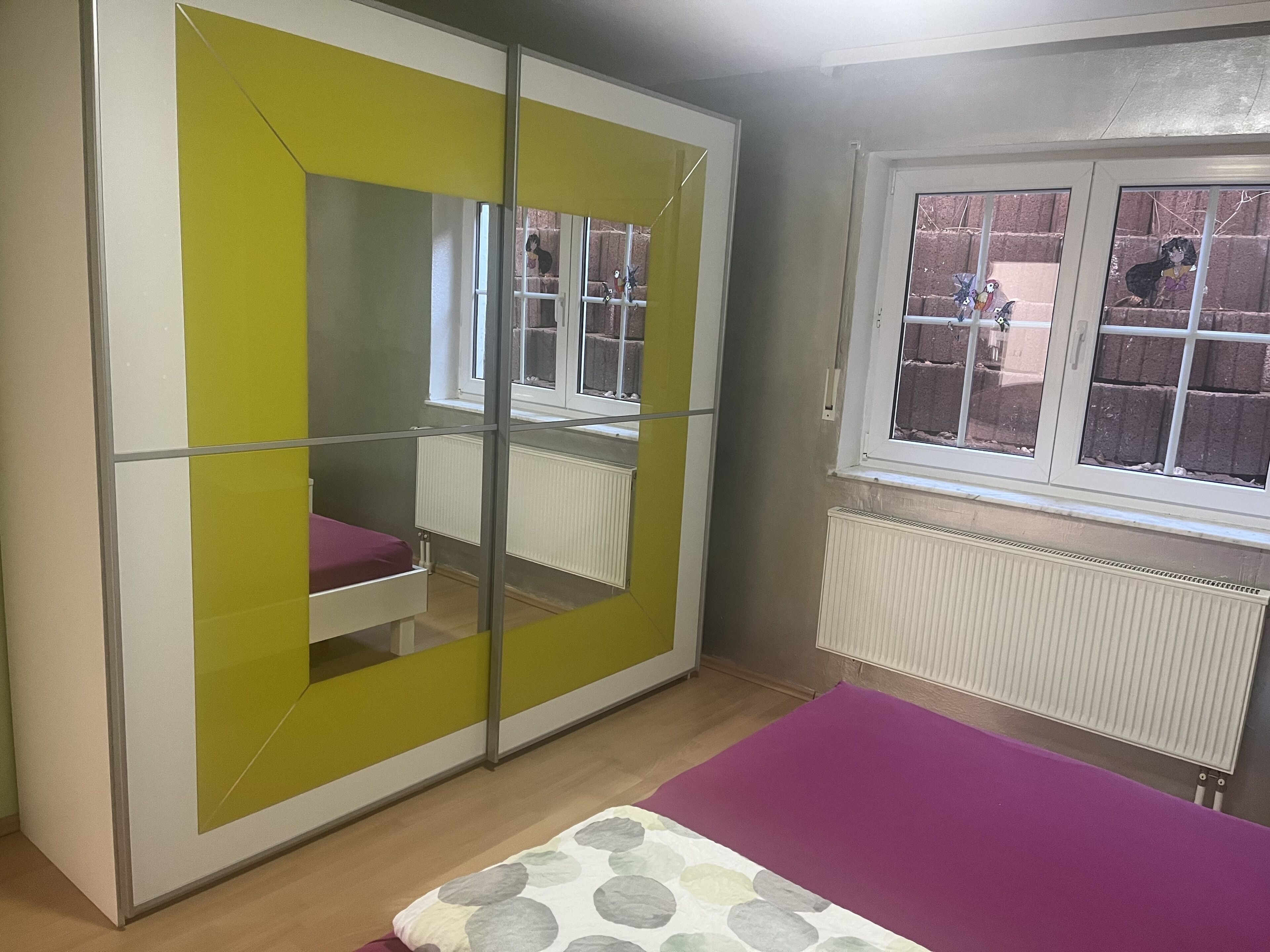 A room with a bed, a wardrobe with a mirror, a window with a view of a brick wall, and a radiator.