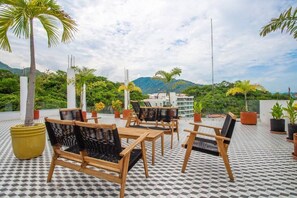 Relax in a serene outdoor space with a geometric patterned floor, wooden tables, and comfortable seating, surrounded by lush greenery and breathtaking mountain views.