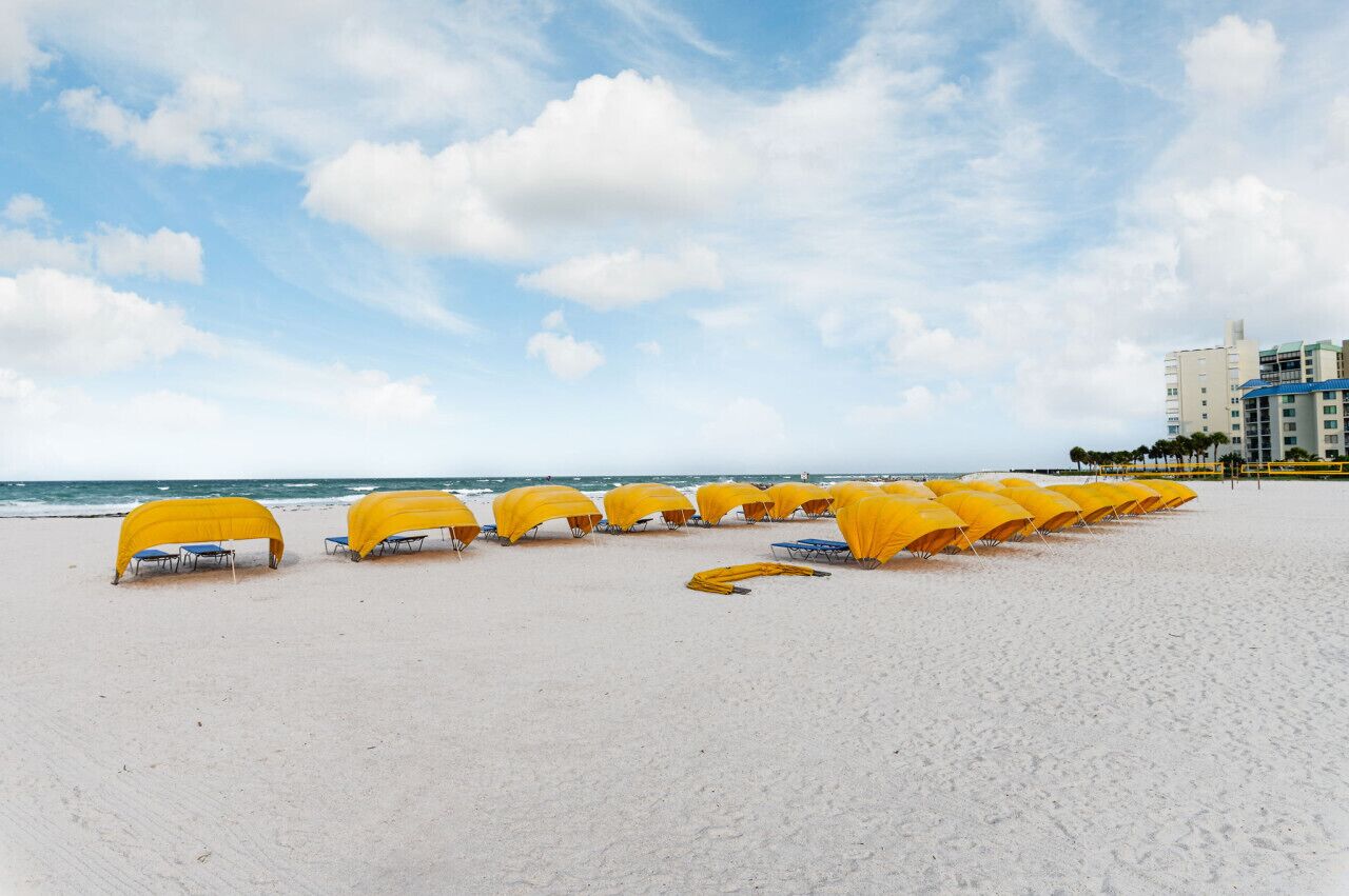 A beach with rows of yellow beach umbrellas and lounge chairs, a clear sky, and a building in the background.