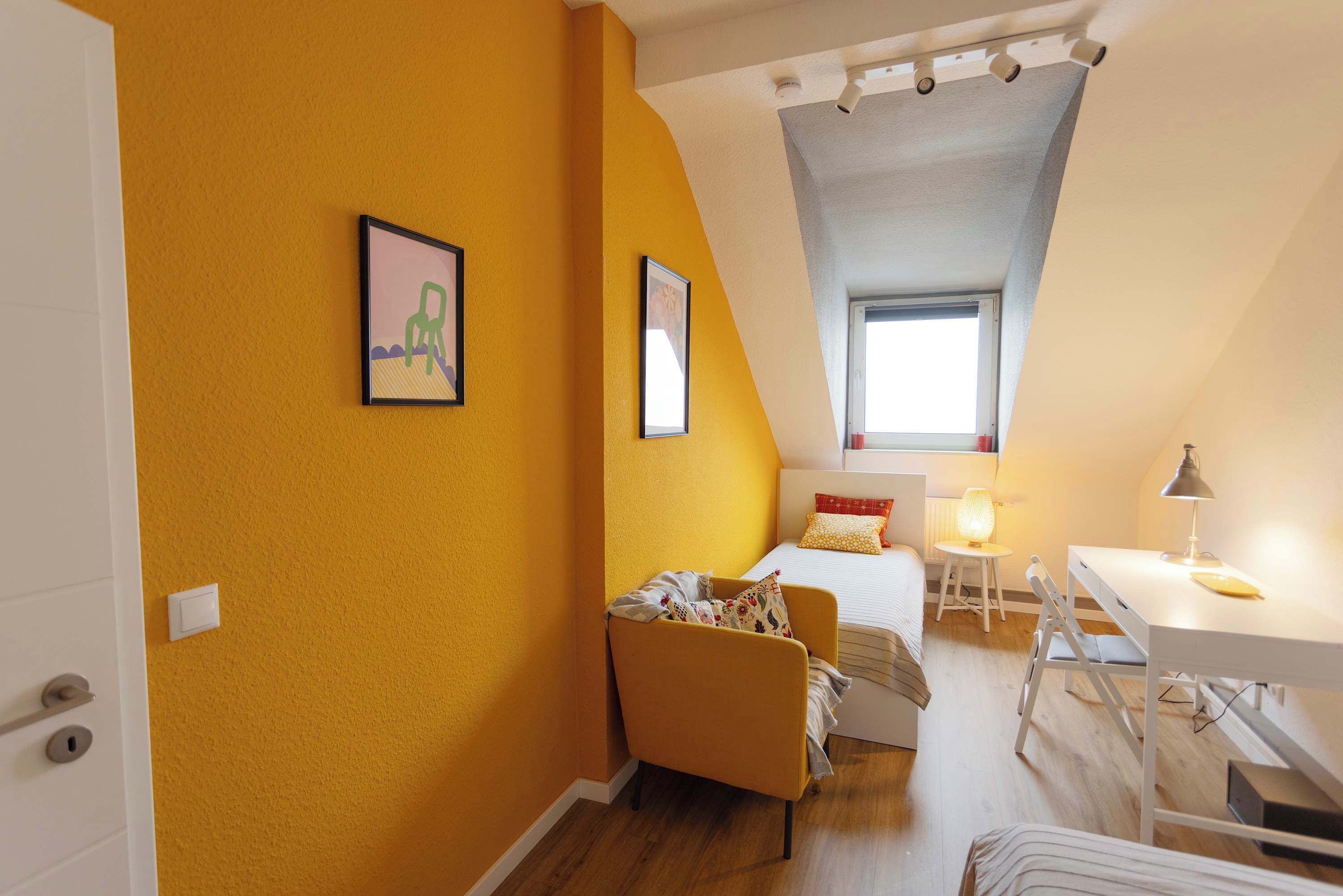 A small, bright room with a yellow wall, a bed, a desk, and a chair.