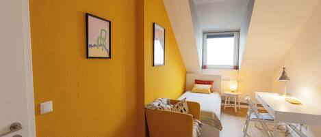 A small, bright room with a yellow wall, a bed, a desk, and a chair.