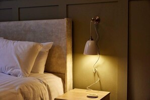 Una cama con sábanas blancas, una mesita de noche con lámpara y un panel en la pared.