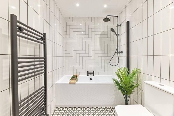 Een moderne badkamer met een visgraatmotief tegels, een vrijstaand bad, een douchekop en een potplant.