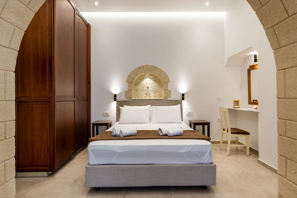 A bedroom with a large bed, wooden wardrobe, and a stone wall arch.