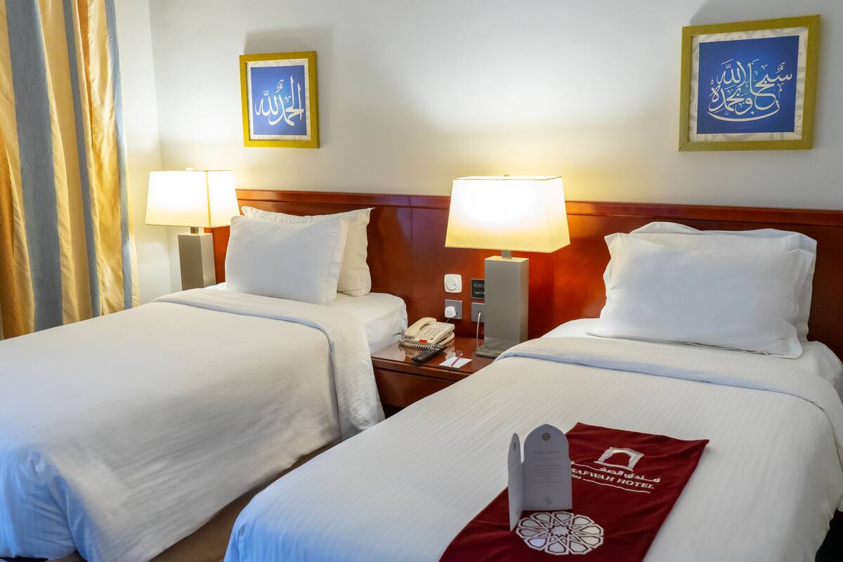 Habitación de hotel con dos camas, cada una con ropa de cama y almohadas blancas, una mesita de noche con lámpara, teléfono y una etiqueta roja con el nombre del hotel.