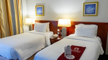 Habitación de hotel con dos camas, cada una con ropa de cama y almohadas blancas, una mesita de noche con lámpara, teléfono y una etiqueta roja con el nombre del hotel.