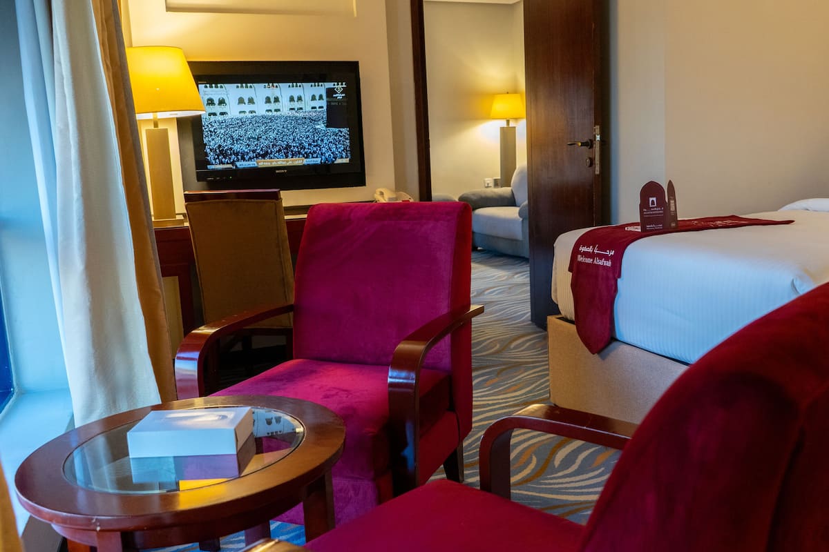 Una habitación de hotel con una cama, un sillón rojo, una mesita, un televisor y una lámpara.