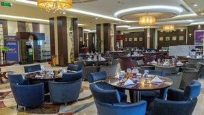 Un hall d’hôtel spacieux, doté de plusieurs espaces salon, notamment des tables rondes et des chaises bleues. De nombreux lustres sont suspendus au plafond, et les murs sont ornés de tableaux encadrés.