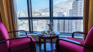Une chambre d’hôtel dotée d’une grande fenêtre donnant sur la ville, de deux fauteuils roses et d’une petite table sur laquelle se trouvent une carafe et des en-cas.