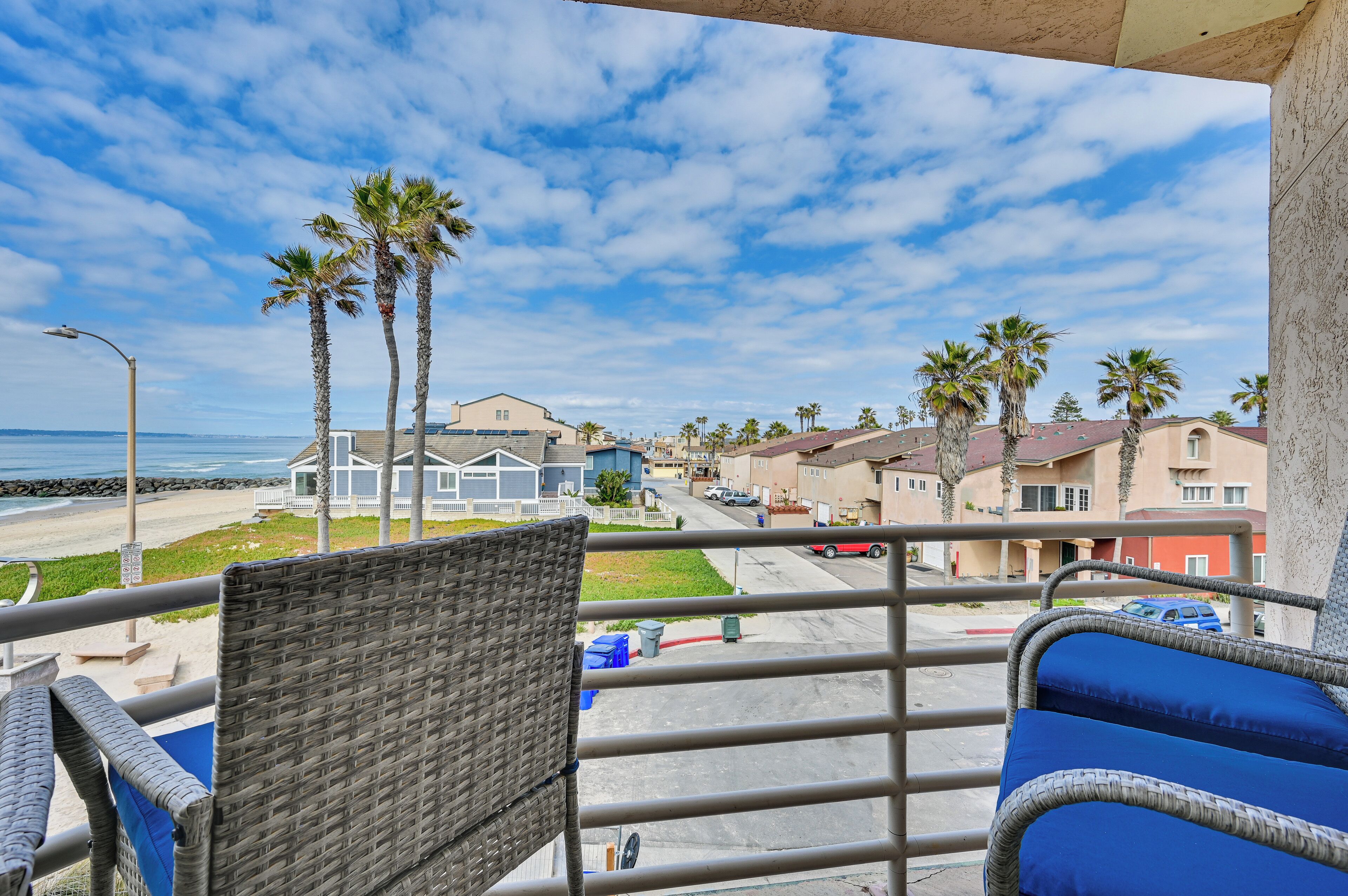 A balcony with wicker chairs and a view of a beach, palm trees, and residential buildings.