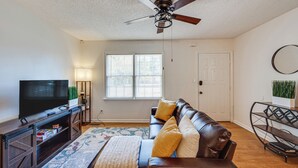 A living room with a ceiling fan, a TV on a wooden stand, a leather sofa with cushions, a window with blinds, and a small side table with a plant. - Cozy North Carolina Retreat Near Fishing & Boating (Washington)
