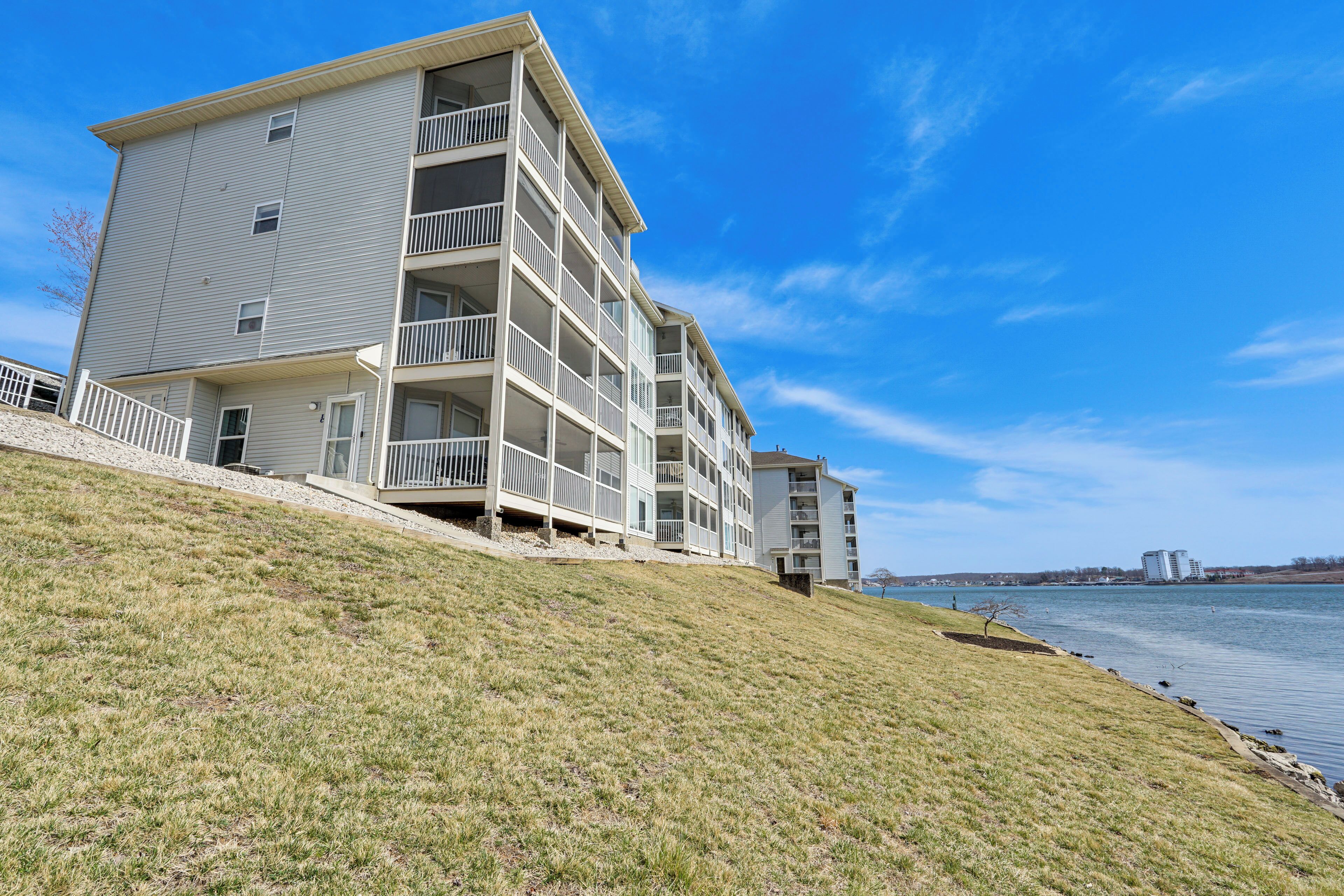 A multi-story building with balconies, situated on a grassy slope overlooking a body of water.
