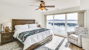 A bedroom with a large bed, a ceiling fan, a view of a bridge, and a chair by the window. - Regatta Bay Condo w/ Balcony + Water Views! (Lake Ozark)