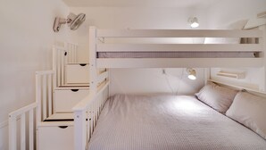 Dormitorio con literas, decoración en blanco, un ventilador de pared y una mesita de noche con lámpara.