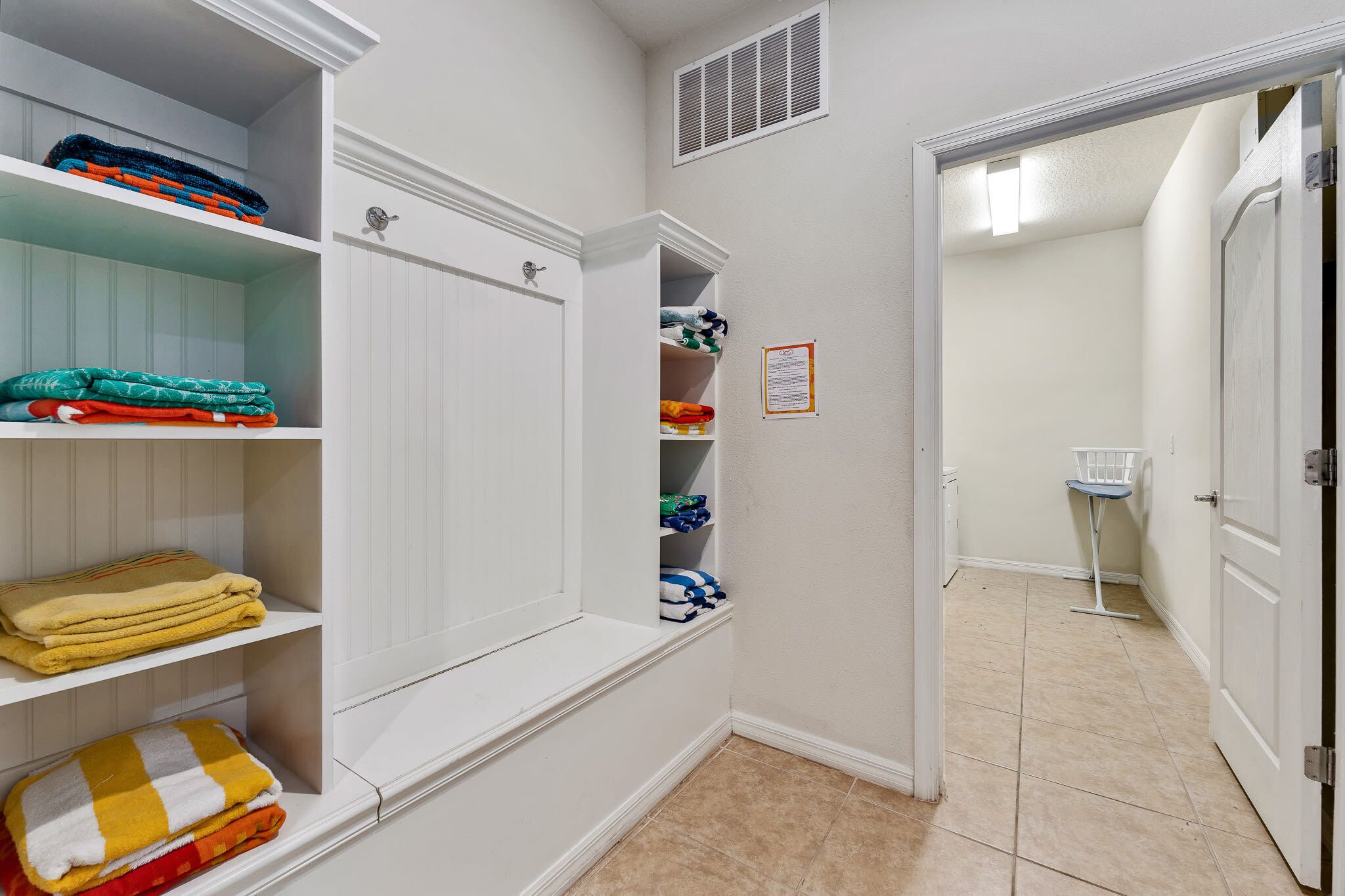 A hallway with white walls, a tiled floor, and a door leading to a bathroom. Shelves are stocked with folded towels and blankets.